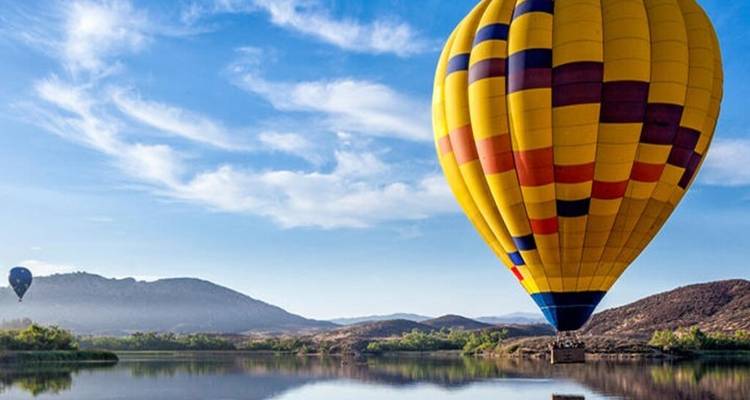 Bright yellow patterned hot-air balloon floating over a calm lake reflecting hills and blue morning sky.
