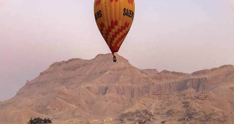 Orange hot-air balloon drifting above rugged desert cliffs and arid landscape at dawn.