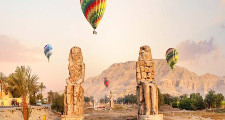 Hot-air balloons drift above the twin seated statues of the Colossi of Memnon with desert mountains in the background under warm early light.