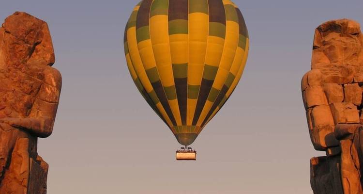 A single yellow-green striped hot-air balloon floats between two ancient statues at dusk, sky tinged with soft light.