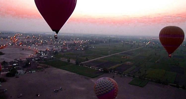 Evening aerial view of farmland and village lights with several colorful balloons rising against a pink horizon.