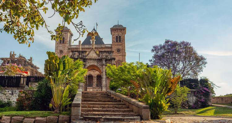 Stone royal palace with decorative towers and lush landscaped stairs under blue sky in Antananarivo.