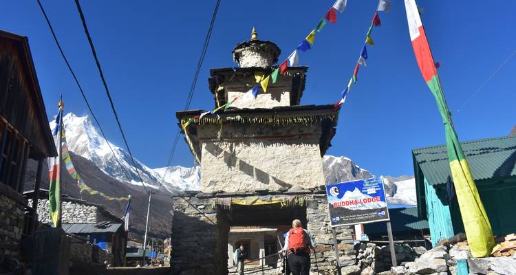 Un trekker se acerca a un chorten en un pueblo de montaña adornado con banderas de oración ondeantes y enmarcado por picos nevados.
