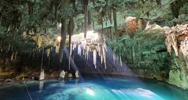 Sonnenstrahlen durchdringen Stalaktiten und fallen in das türkisfarbene Wasser einer unterirdischen Cenote-Höhle.