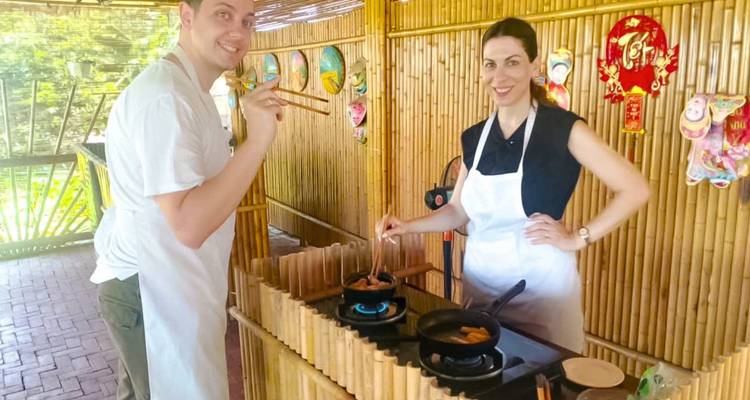 Travellers wearing aprons cook spring rolls on gas burners inside a bamboo kitchen during a Vietnamese class.