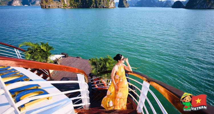 Woman in flowing yellow dress gazes over emerald waters and limestone spires from a cruise ship railing.