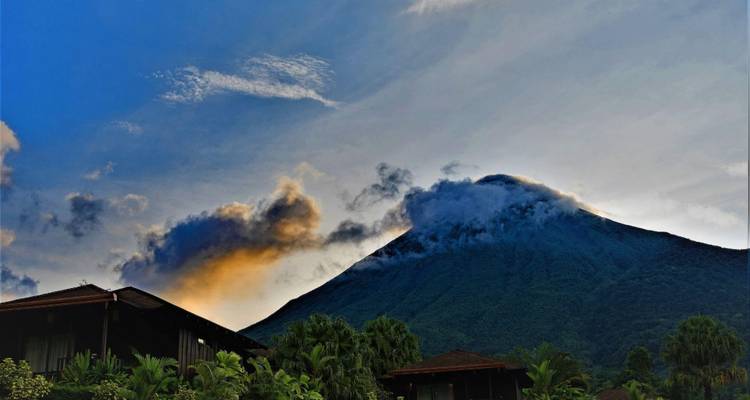 Volcano peak shrouded in cloud with warm sunset light and silhouette of buildings below.