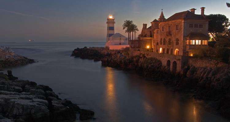 Leuchtturm und Herrenhaus auf einer felsigen Bucht nahe Cascais, die in der Abenddämmerung erleuchtet sind und sich in ruhigen Gewässern spiegeln.