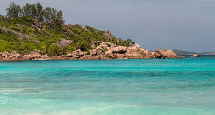 Turquoise sea crashes softly against smooth granite boulders fringed by tropical forest.
