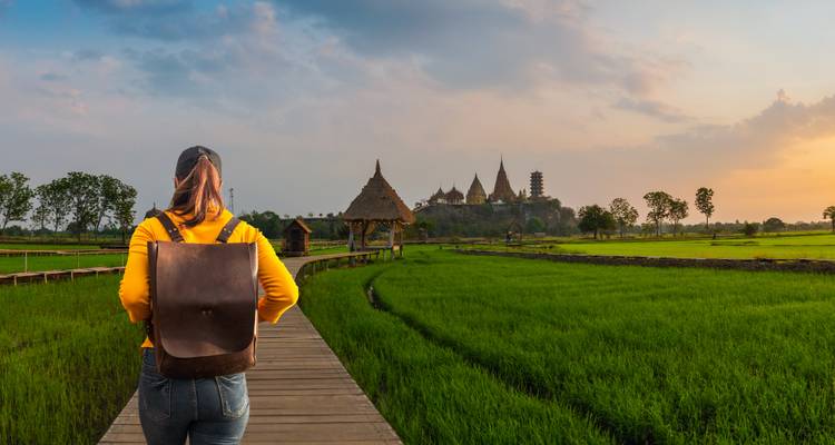 Achteraanzicht van reiziger met rugzak die over een houten pad loopt door groene rijstvelden naar een tempel op de heuvel.