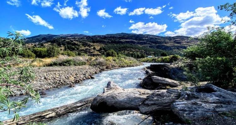 A fast-flowing mountain stream tumbles past rocky banks under a bright blue sky.