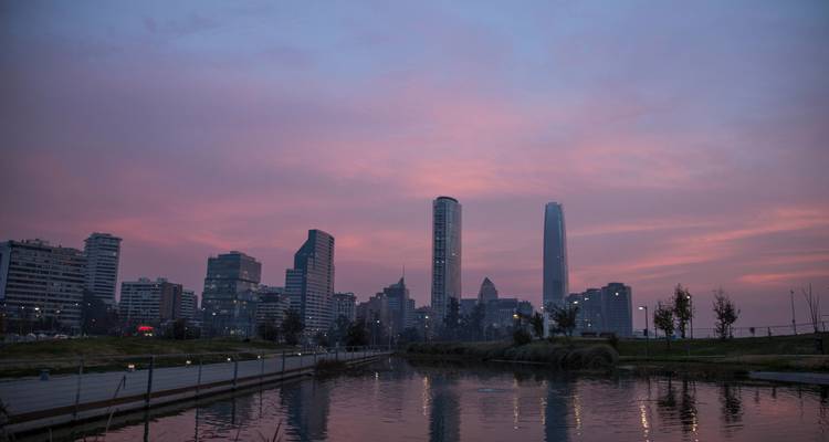 A modern skyline with high-rise towers is reflected in a calm pond at sunset.