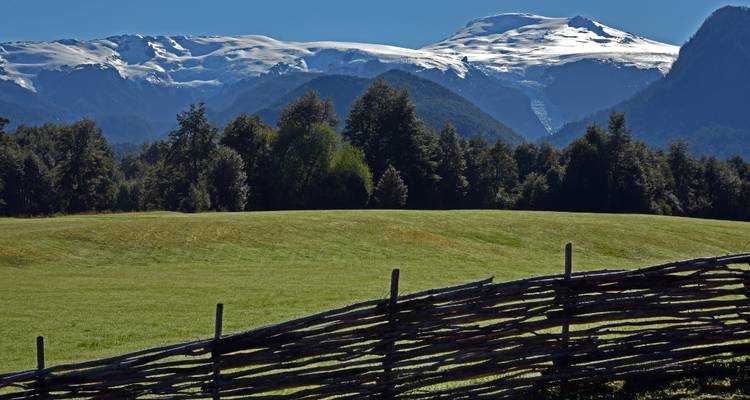 A green meadow bordered by a rustic wooden fence with glaciated peaks beyond.
