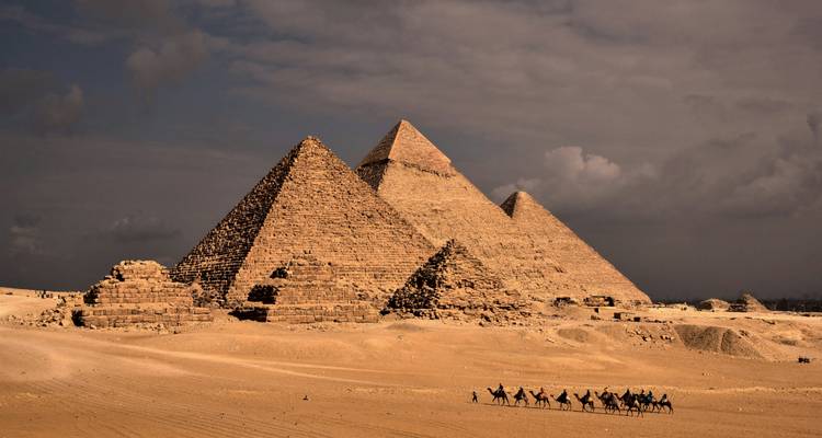The Great Pyramids of Giza dominate the desert horizon as a camel caravan passes below heavy evening clouds.
