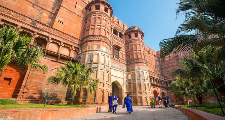 Toeristen in kleurrijke outfits verkennen een ingewikkeld gedeelte van het Agra Fort dat versierd is met Mughal-architectuur en palmbomen.