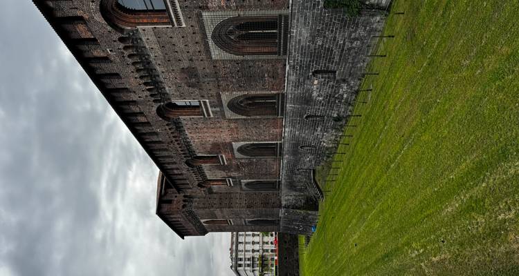 A long brick fortress wall with arched windows overlooks a neatly mown grassy moat under an overcast sky.