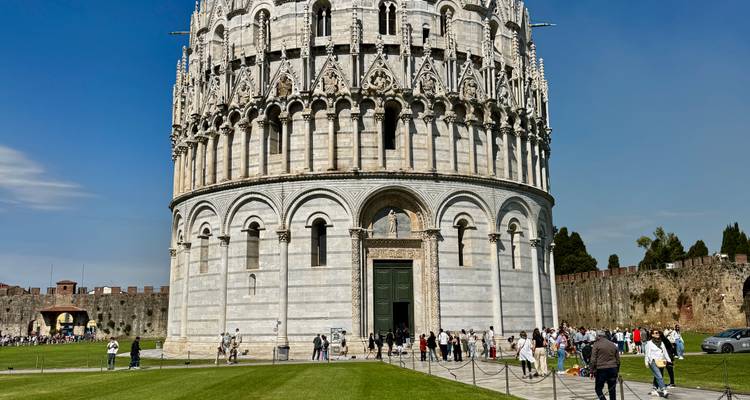 The Pisa Baptistery with tourists gathered on the surrounding lawn under a clear blue sky.
