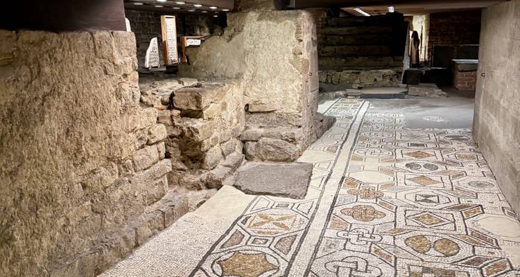 Excavated underground section with ancient stone walls and mosaic tile flooring lit by modern lights.