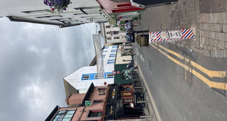 Narrow city street lined with colorful pubs and shops under an overcast sky with a few pedestrians walking along the road.
