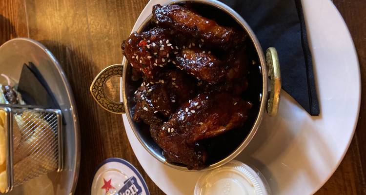 Close-up of glazed chicken wings sprinkled with sesame seeds served in a metal bowl at a wooden pub table.