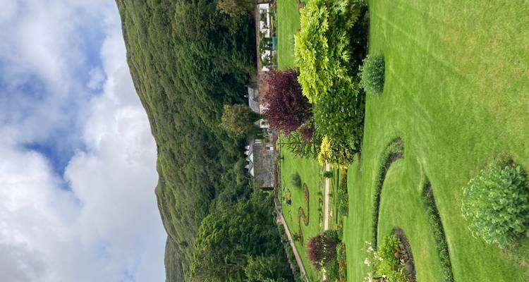 Manicured garden with curved flower beds set against green hills and scattered cottages under partly cloudy skies.