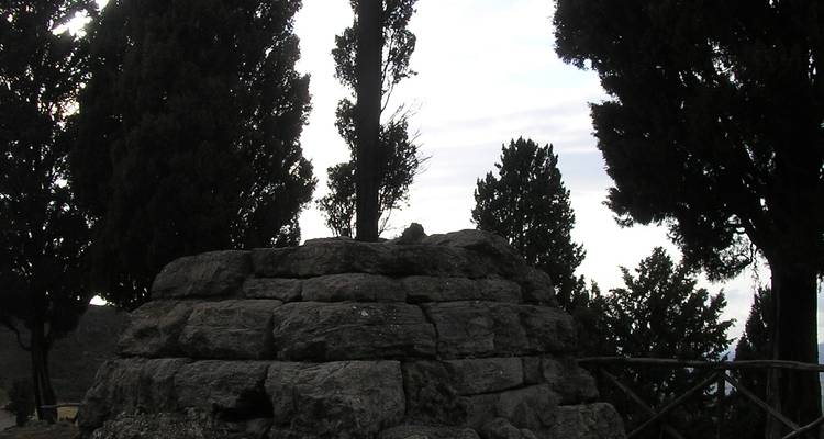 Silhouette sombre d'un cairn de pierre entouré de grands cyprès sous un ciel nuageux.
