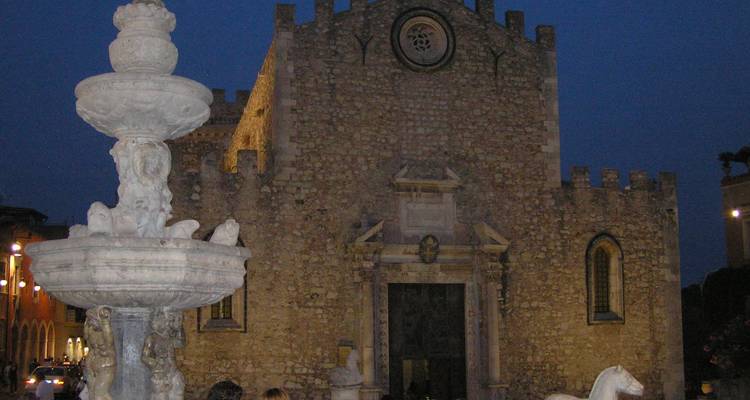 Vue nocturne de la cathédrale en pierre de Taormina avec une fontaine ornée au premier plan.