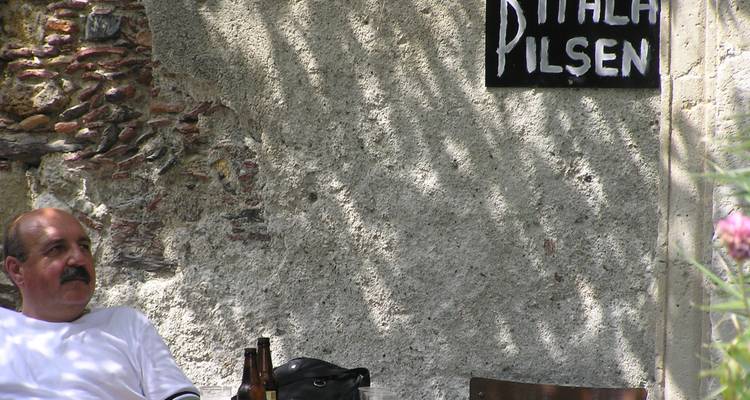 Un homme se détendant avec une bière sous la lumière du soleil tamisée à côté d'un mur rustique annonçant une pilsner locale.