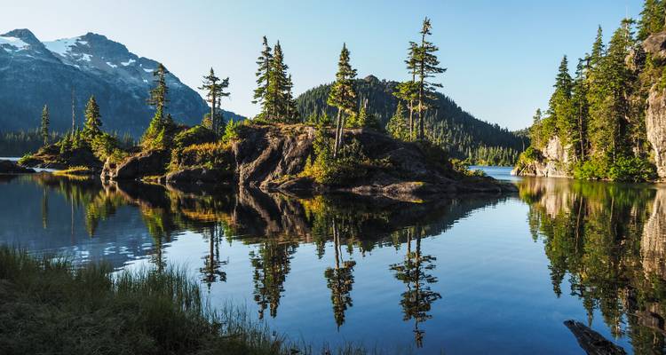 Crystal-clear alpine lake with reflections of pine-clad islets and snow-capped peaks in early morning light.