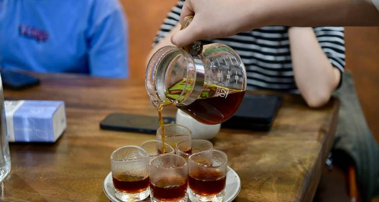 Hand pours brewed coffee from a glass decanter into small tasting glasses on a plate.