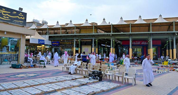 Drukke buitenmarkt met mannen in witte thobes die onder getende daken langs kramen struinen.