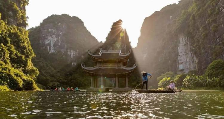 Rowboat passing ornate riverside pagoda framed by karst mountains with sunburst behind peak.