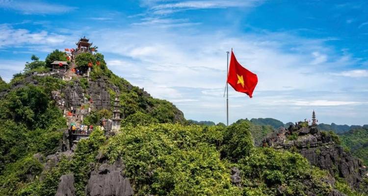 Vietnamese flag flying atop rocky hill with pagoda and distant karst landscape under blue sky.