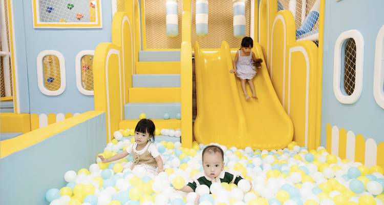 Three small children playing in colorful indoor ball pit with yellow slide.
