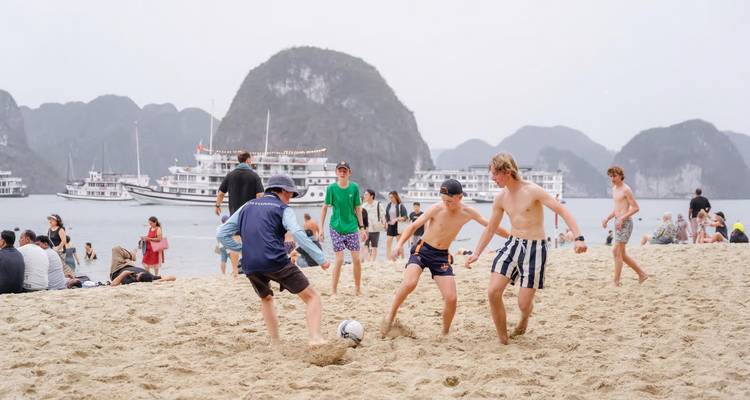 Teens and travellers playing beach football with Halong Bay karst towers behind.