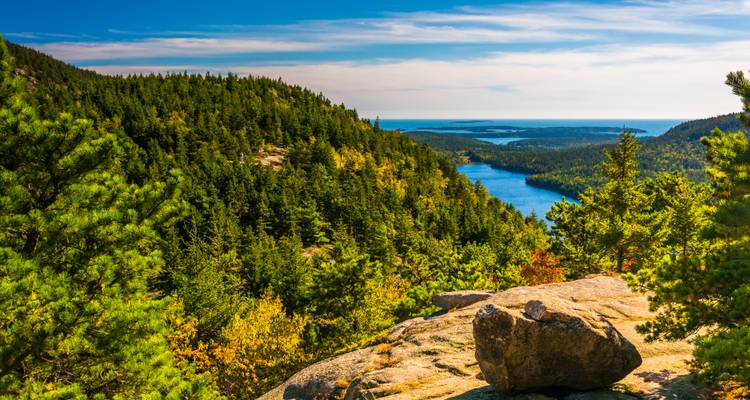 Panoramic view over evergreen forests, a long blue lake, and distant sea from a granite summit in Acadia.