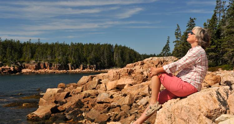 An older woman relaxes on sunlit pink granite rocks beside a calm coastal inlet lined with evergreen forest under a clear blue sky.