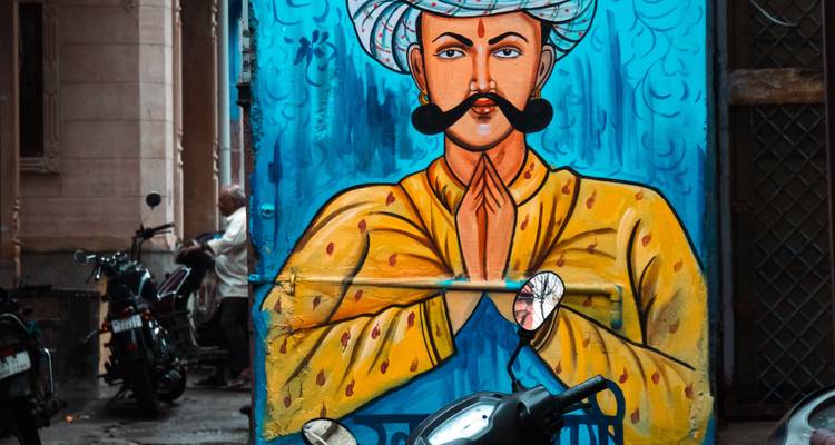 Kleurrijke street-art muurschildering van een man met een snor en gevouwen handen op een blauwe muur.