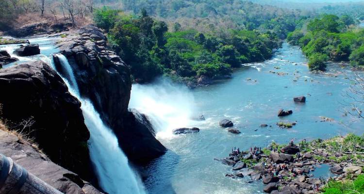 Large waterfall plunges into a broad river surrounded by lush forested hills, visitors gather on rocks below.
