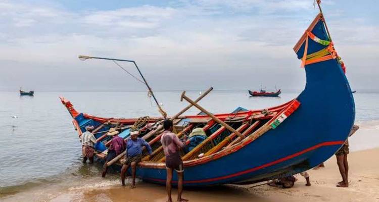 Fishermen push a brightly painted blue wooden boat into calm sea waters on a sandy beach.