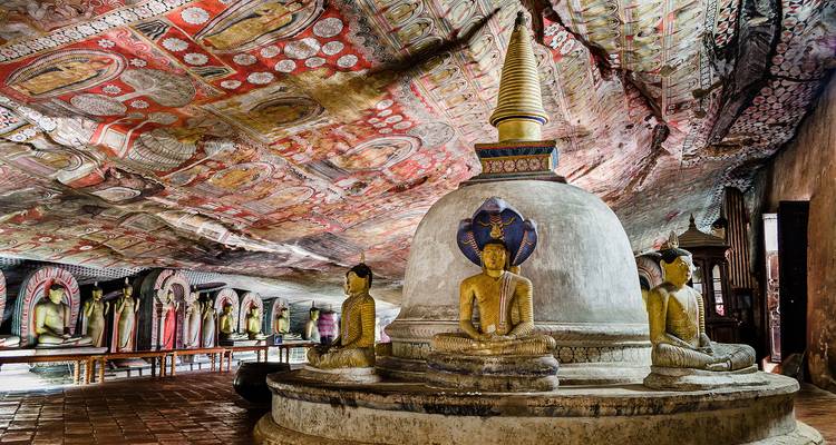 Intricately painted cave temple interior with a central stupa surrounded by Buddha statues under a patterned ceiling.