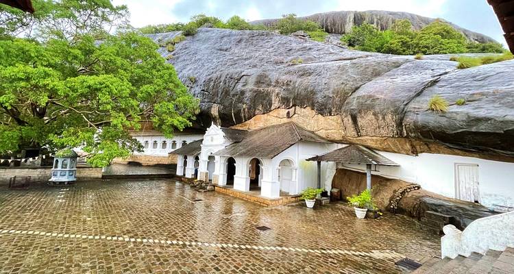 Rock-sheltered white temple buildings beside a cobbled courtyard beneath a huge granite overhang.