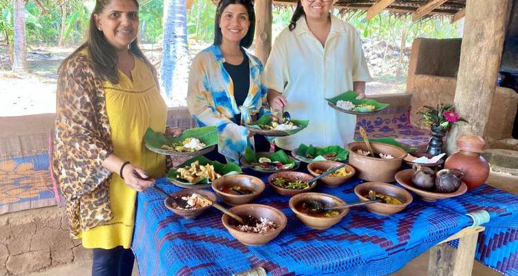 Three women standing behind a rustic table of traditional Sri Lankan dishes inside an open-air kitchen.