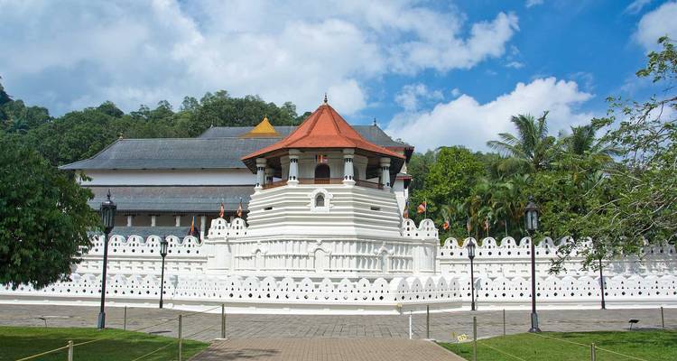 White Temple of the Tooth Relic with red roof and moat set against green trees and blue sky.