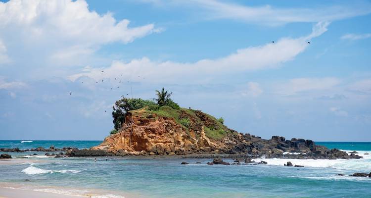 Small rocky island capped with greenery just off a tropical shoreline in turquoise waters.