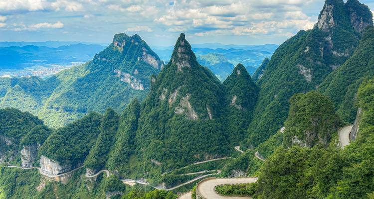 Vue panoramique de pics escarpés et verdoyants ainsi qu'une route de montagne sinueuse sous un ciel partiellement nuageux