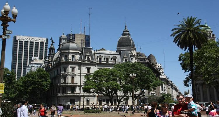 Historic European-style buildings and palm tree ring a busy Buenos Aires plaza filled with pedestrians under blue sky.