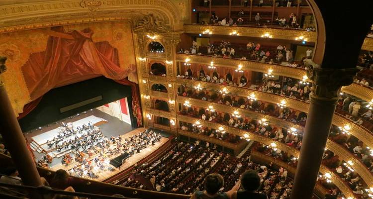 Ornate interior of Teatro Colón with glowing chandeliers, gilded balconies and an orchestra performing on stage.