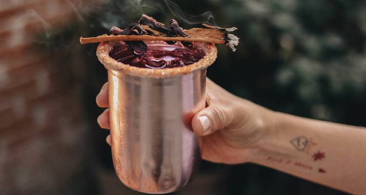 Hand hält einen dekorativen handwerklichen Cocktail in einem metallischen Becher mit rauchigem Hibiskus-Aroma.