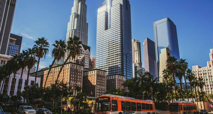 Modern skyscrapers and palm trees rise above a busy downtown Los Angeles street with an orange city bus in the foreground.